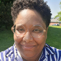This is a picture of a brown-skinned person with glasses and dreadlocks, looking straight at the camera and smiling.