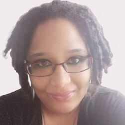 This is a picture of a brown-skinned person with glasses and dreadlocks, looking straight at the camera and smiling.